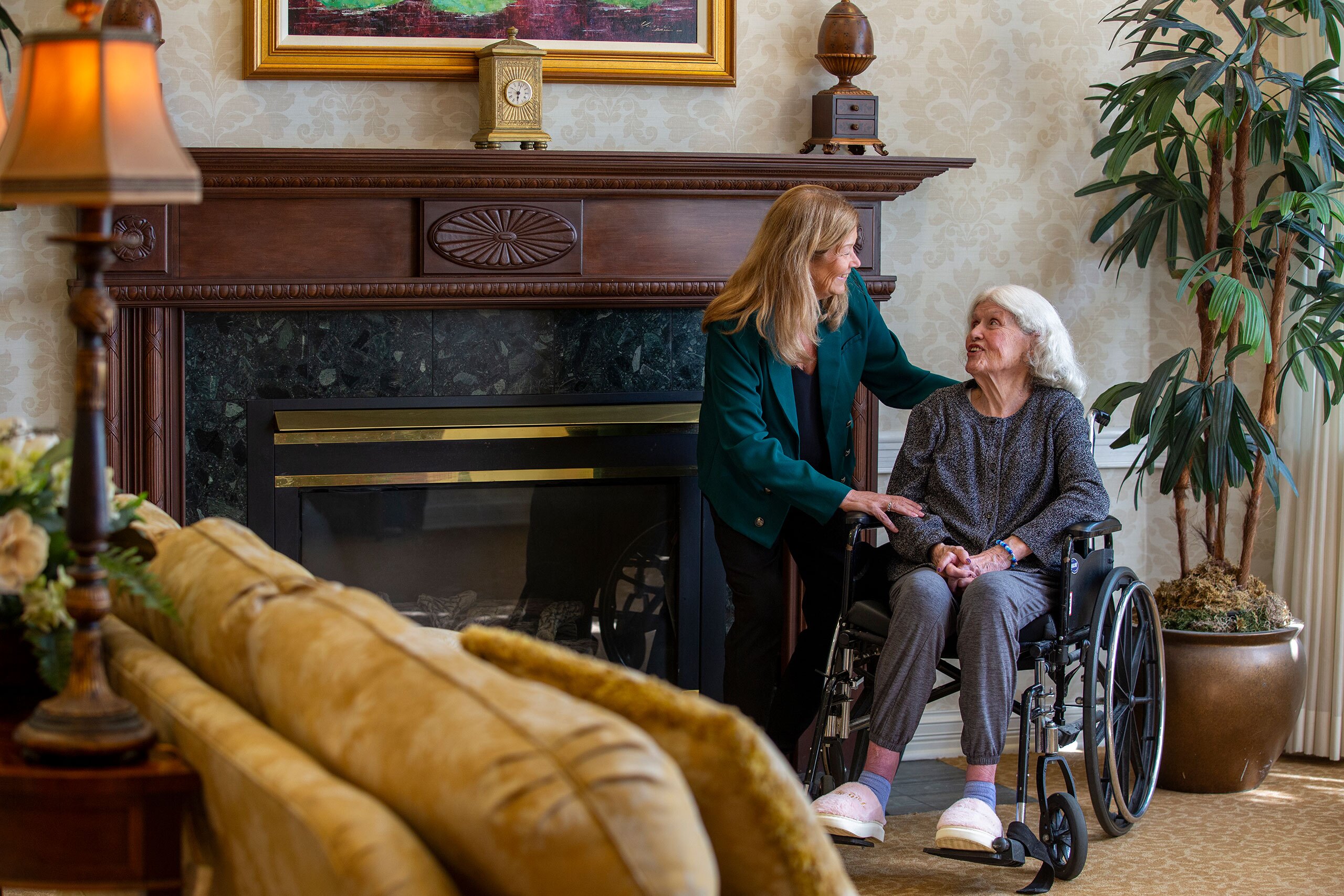 A resident sits in a wheelchair speaking to another lady in front a fireplace.