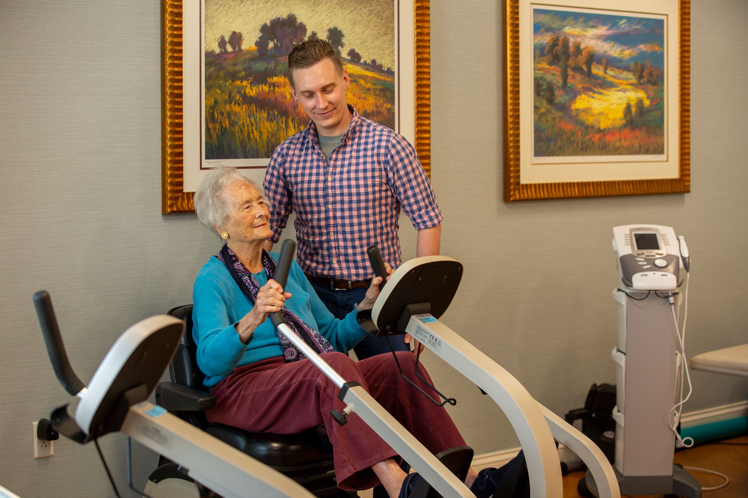 A resident sits in a therapy machine holding onto handlebars while a younger man watches on.