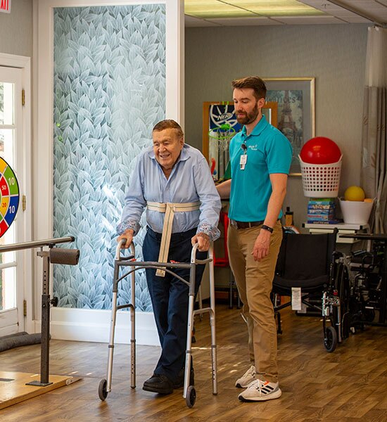 A resident in the walks with a walker while being supported by staff.