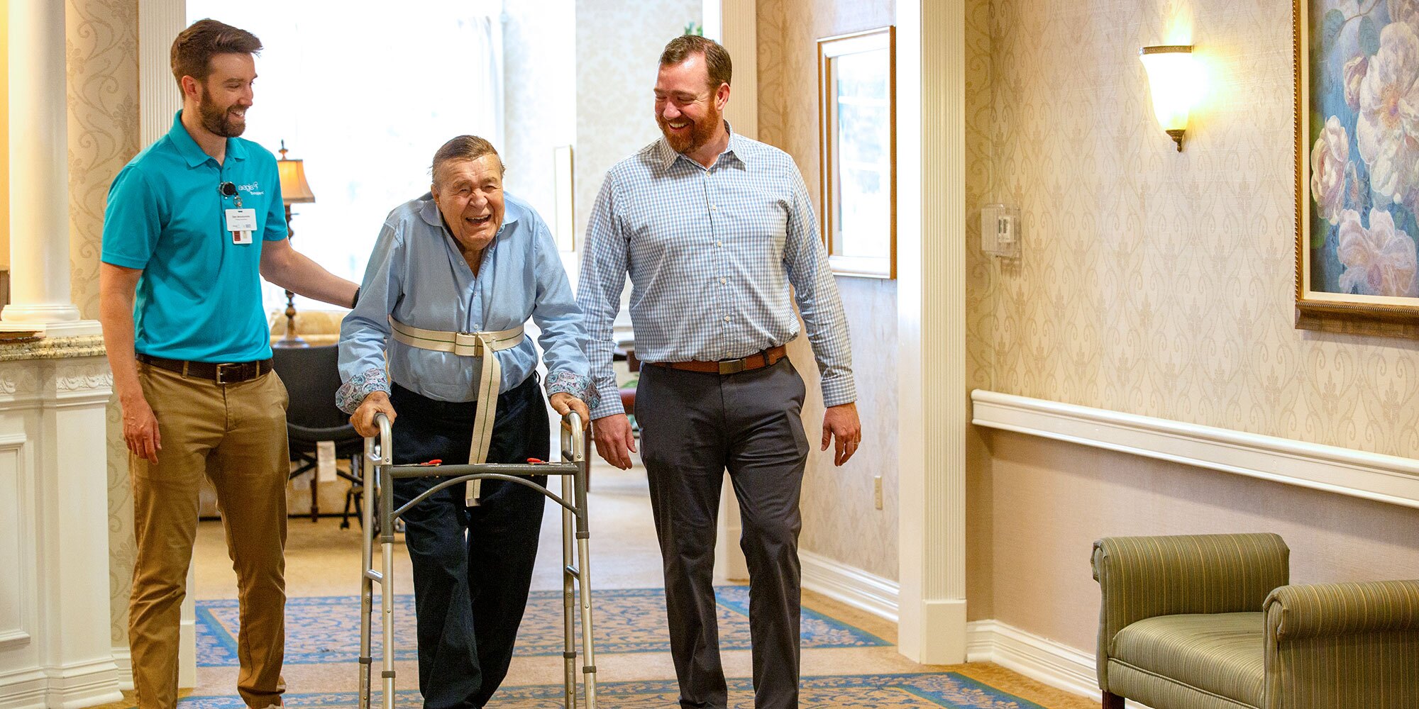 A man in a walker walks down a hallway with support staff walking along with him. He smiles.