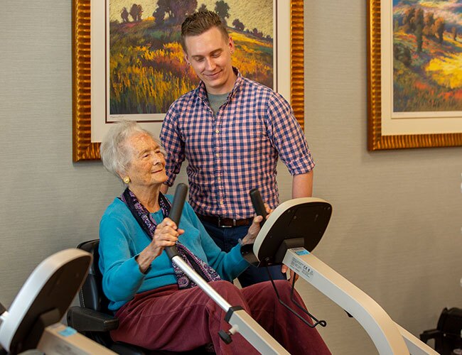 A resident sits in a therapy machine holding onto handlebars while a younger man watches on.