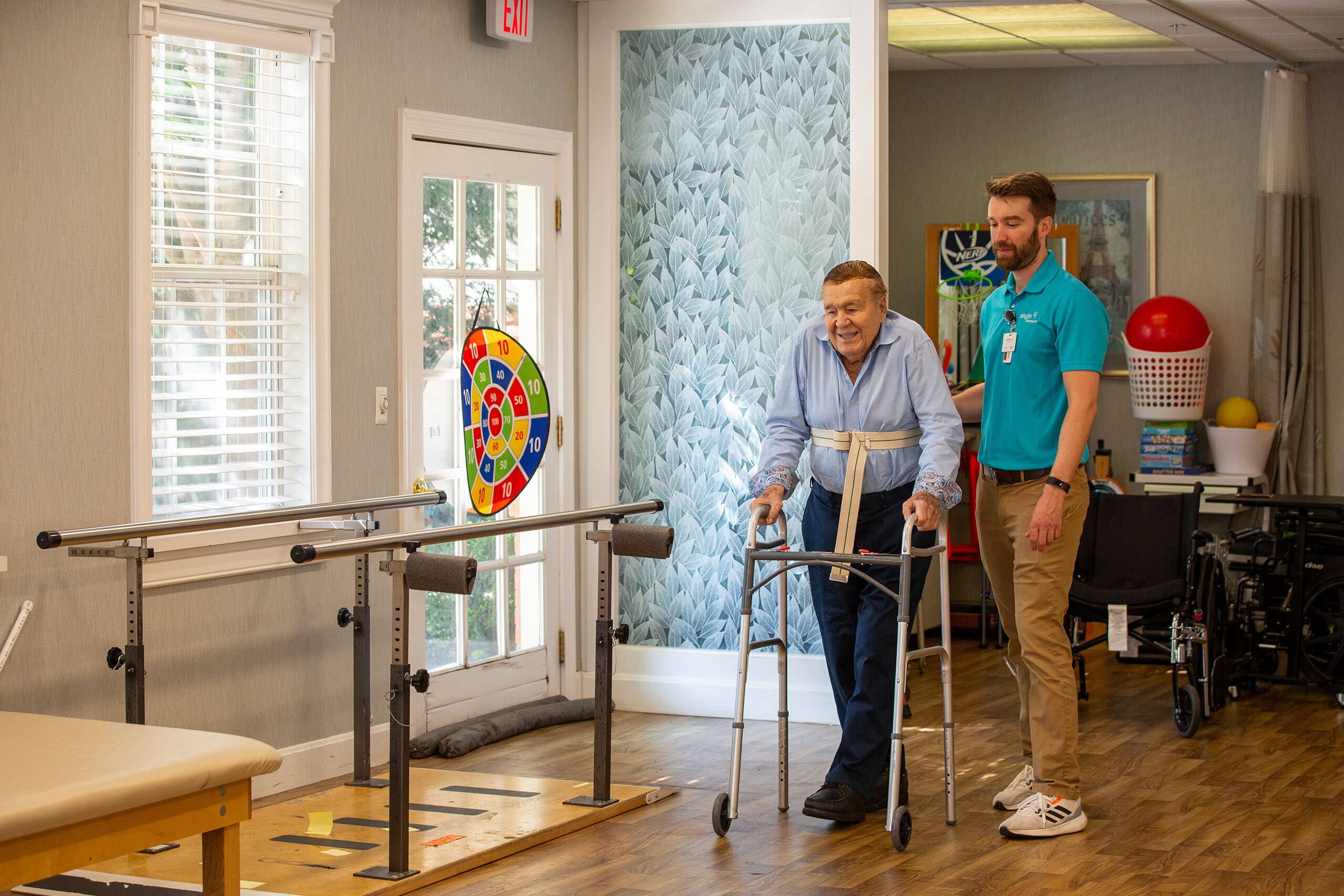 A resident is supported by a therapy staff member while walking with a walker.
