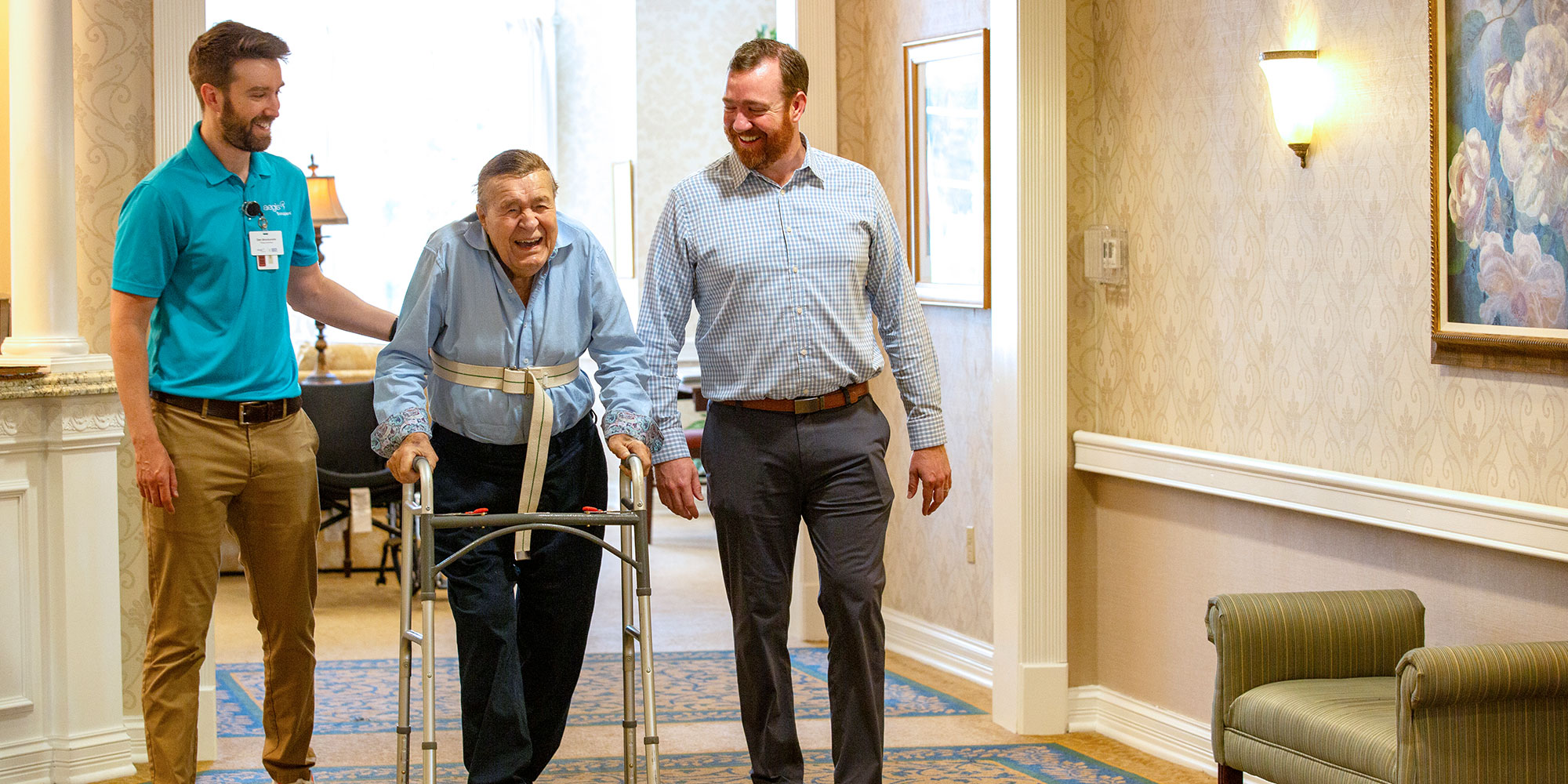 A man in a walker walks down a hallway with support staff walking along with him. He smiles.